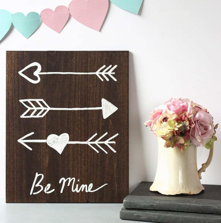 This cute wooden sign with arrows and hearts is an easy and rustic addition to your valentines day decor. It's displayed here under a heart banner and next to a white vase of flowers. 