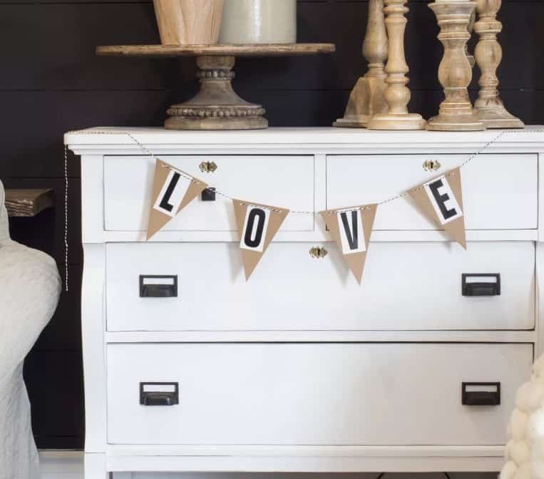 This Valentine's decoration features a simple pennant spelling out the word love on basic paper triangles. The photo shows it hung in front of a white dresser. 