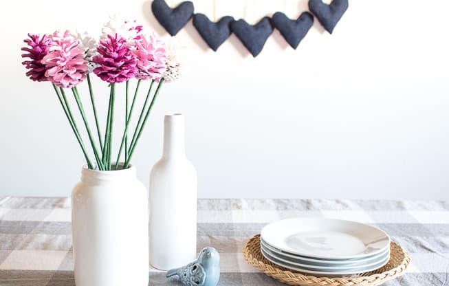 Pine cones painted in various shades of pink are turned into lovely faux flowers displayed on a table in a white mason jar