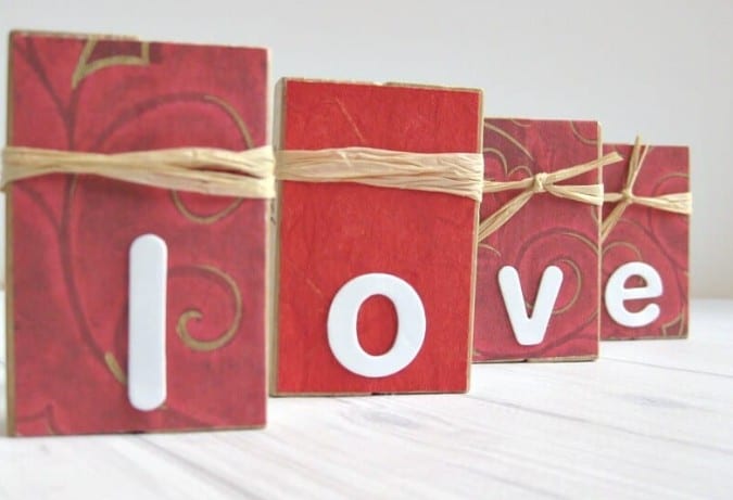 Valentine’s Décor with Upcycled Game Blocks: 4 sets of wooden game blocks covered in red paper, wrapped in straw. They display the letters L.O.V.E.
