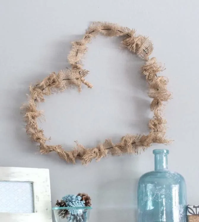 A wire heart wrapped in burlap is a fun farmhouse variation on traditional valentines day decorations. Here it is displayed on a wall above a vase and a picture frame. 