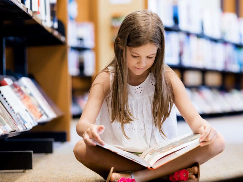 It's easier to homeschool without curriculum when you spend lots of time in the library. In this picture, a young girl sits on the floor of a library reading a book. 