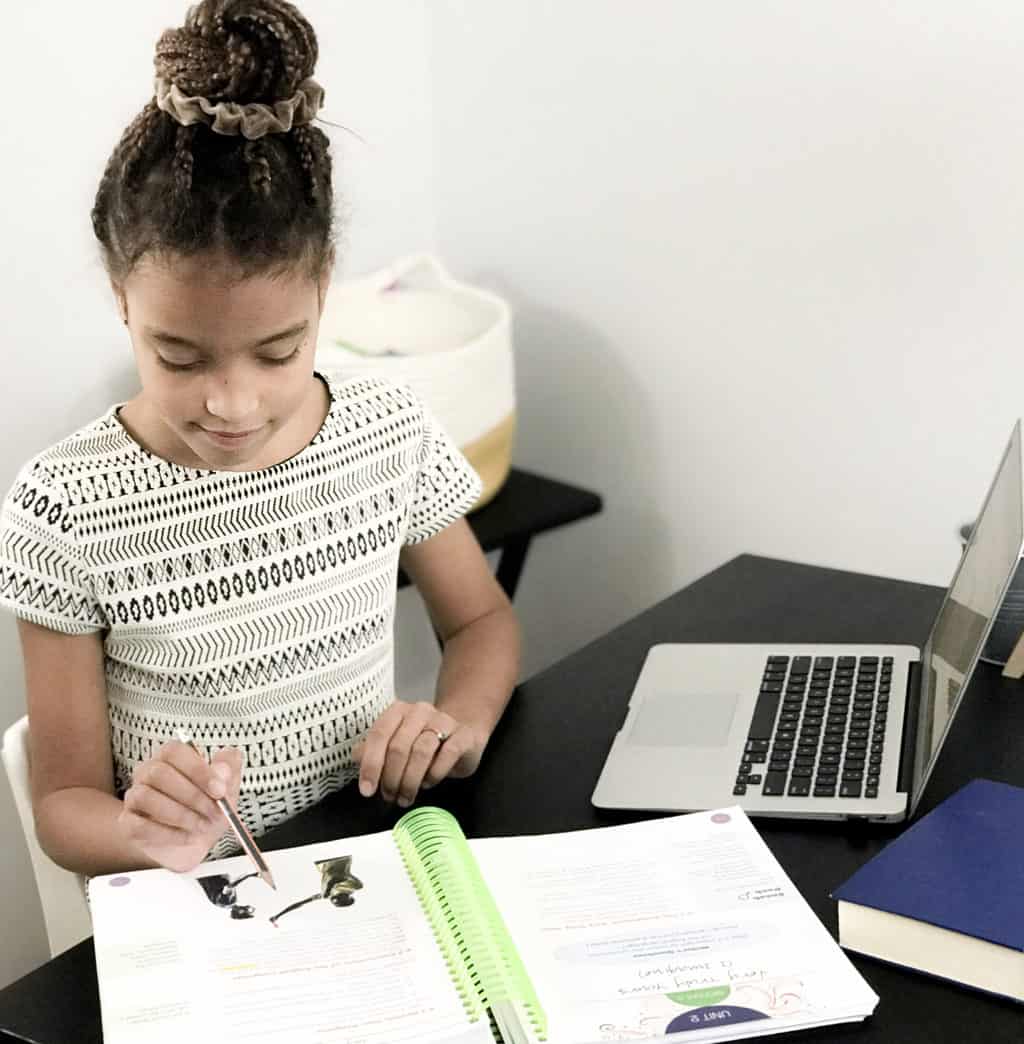 a young girl sitting at a desk writing on a computer and working from the Writers in Residence homeschool writing curriculum book