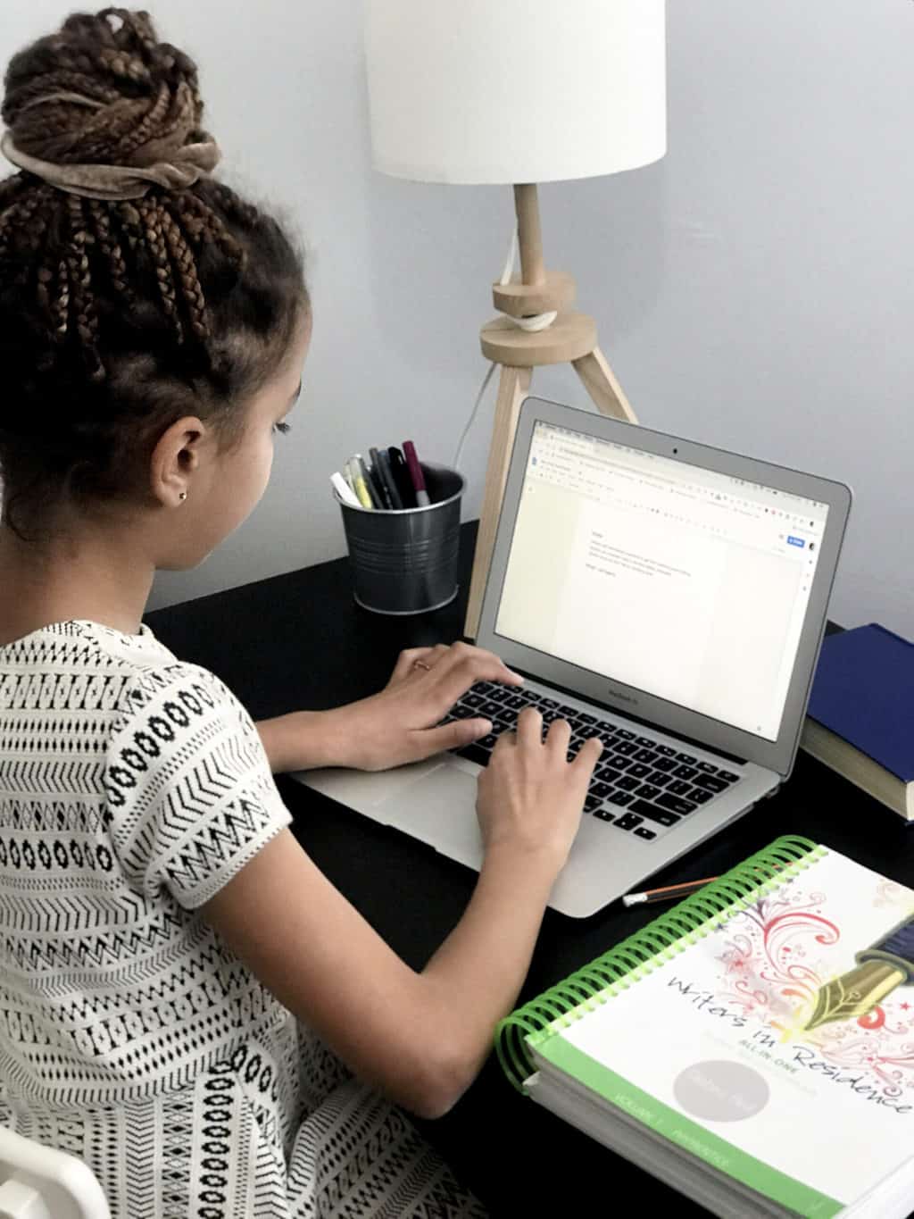 a young girl sitting at a desk writing on a computer and working from the Writers in Residence homeschool writing curriculum book