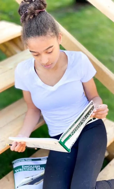 A teenage girl studies a high school English curriculum book while sitting on wooden steps outside