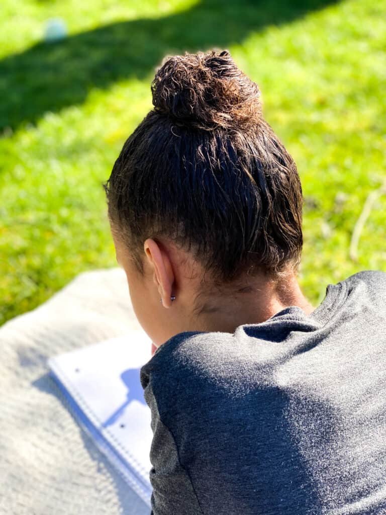 A girl writes in a notebook while lying on the ground outside. The writing portion of the Advanced Writing online high school writing course can be done online or offline. 