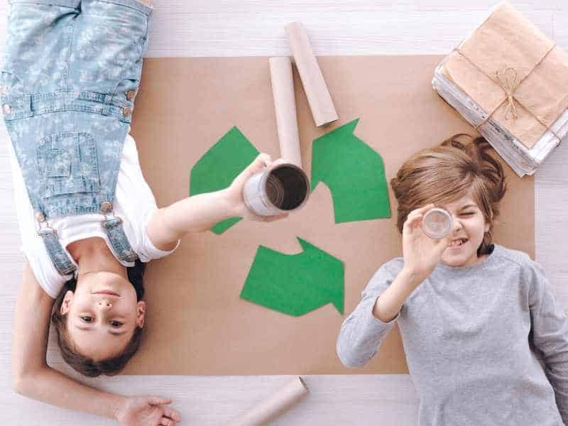Two kids lying on a piece of paper painted with a recycling sign and surrounded by recyclable materials. Household items like these are perfect inputs for our Earth Day Activities for Kids