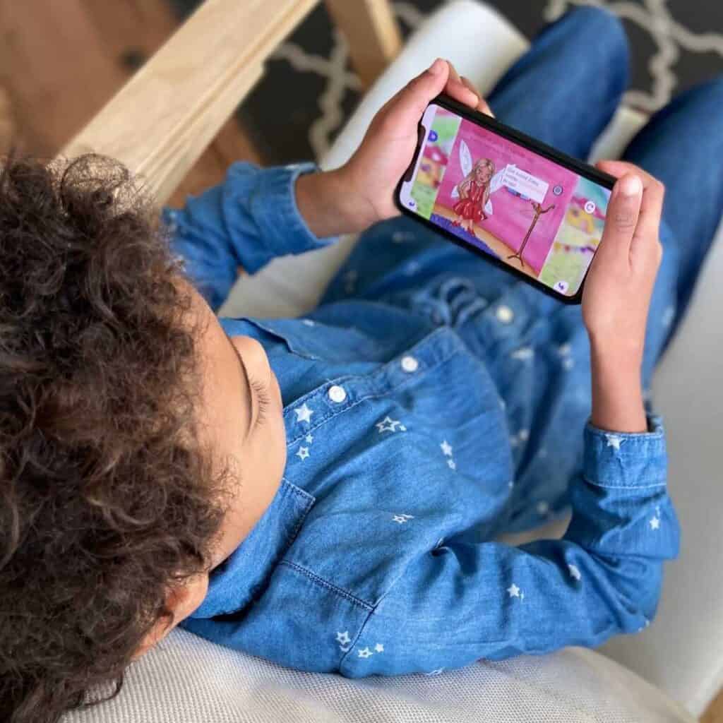 A little girl sitting in a chair playing with a Spanish learning app on a smartphone. Text overlay: Teach Kids Spanish with this supercute app