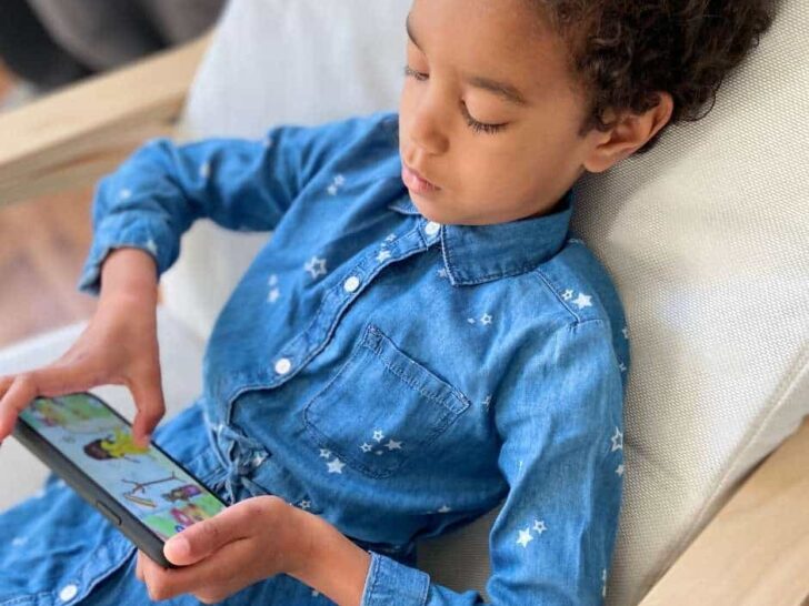 A little girl sitting in a chair playing with a Spanish learning app on a smartphone. Text overlay: Teach Kids Spanish with this supercute app