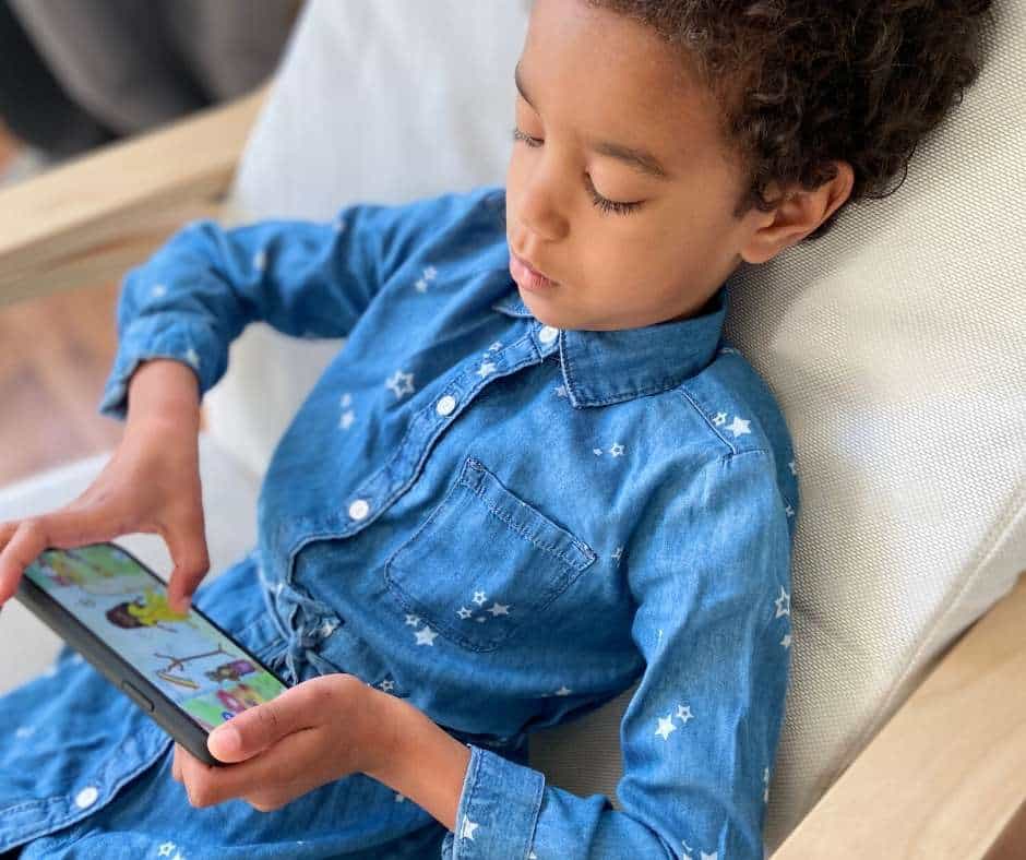 A little girl sitting in a chair playing with a Spanish learning app on a smartphone. Text overlay: Teach Kids Spanish with this supercute app