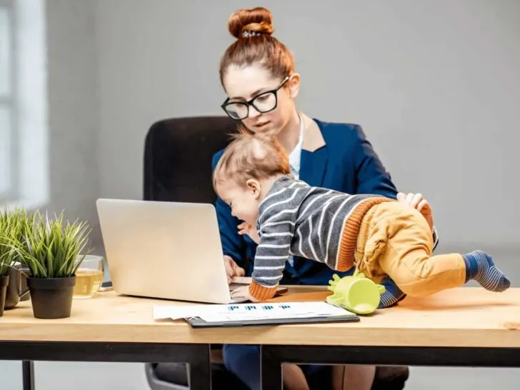 a mom works on a computer while a child crawls on it | learn how to schedule your day as a work at home homeschooler