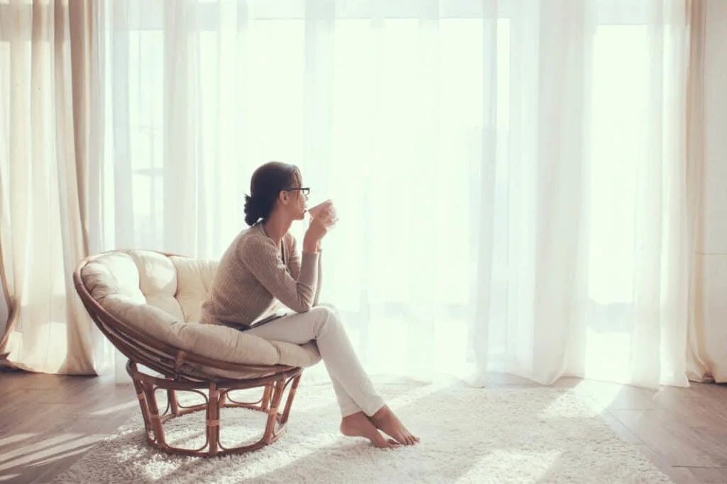 a woman sits in a chair and drinks coffee by a window