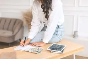 a woman sits at a desk with a journal, some money, and a tablet