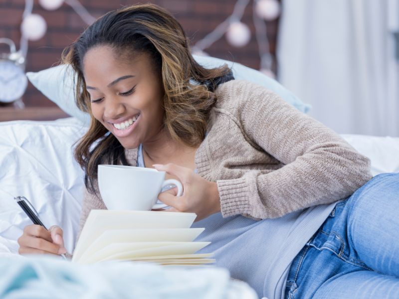 a woman lies on a bed and writes in a gratitude journal while holding a cup