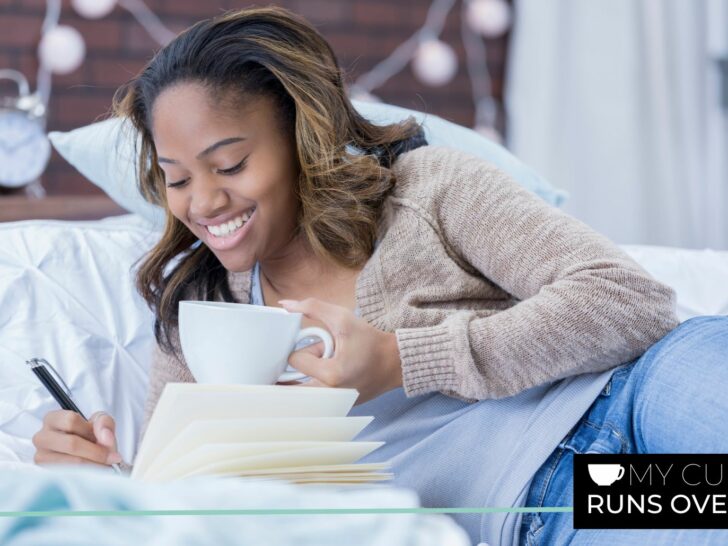 a woman lies on a bed and writes in a gratitude journal while holding a cup